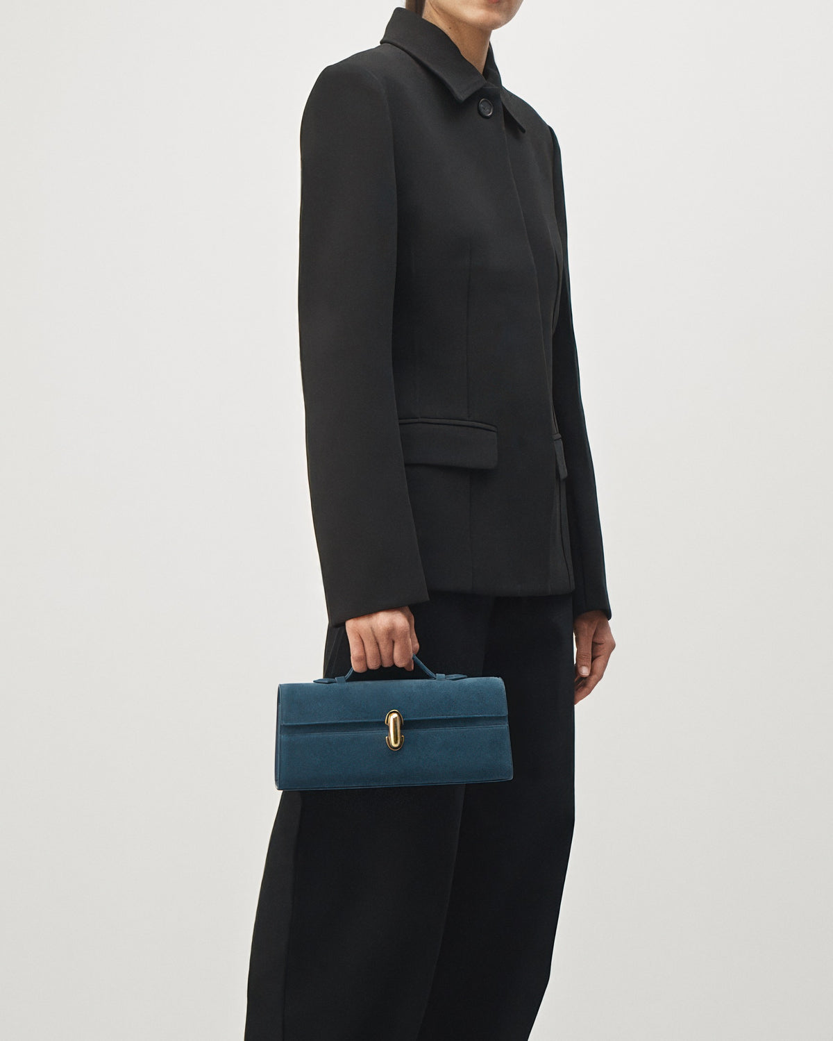 on model view of a baltic blue suede top handle bag with a gold clasp on a white background 