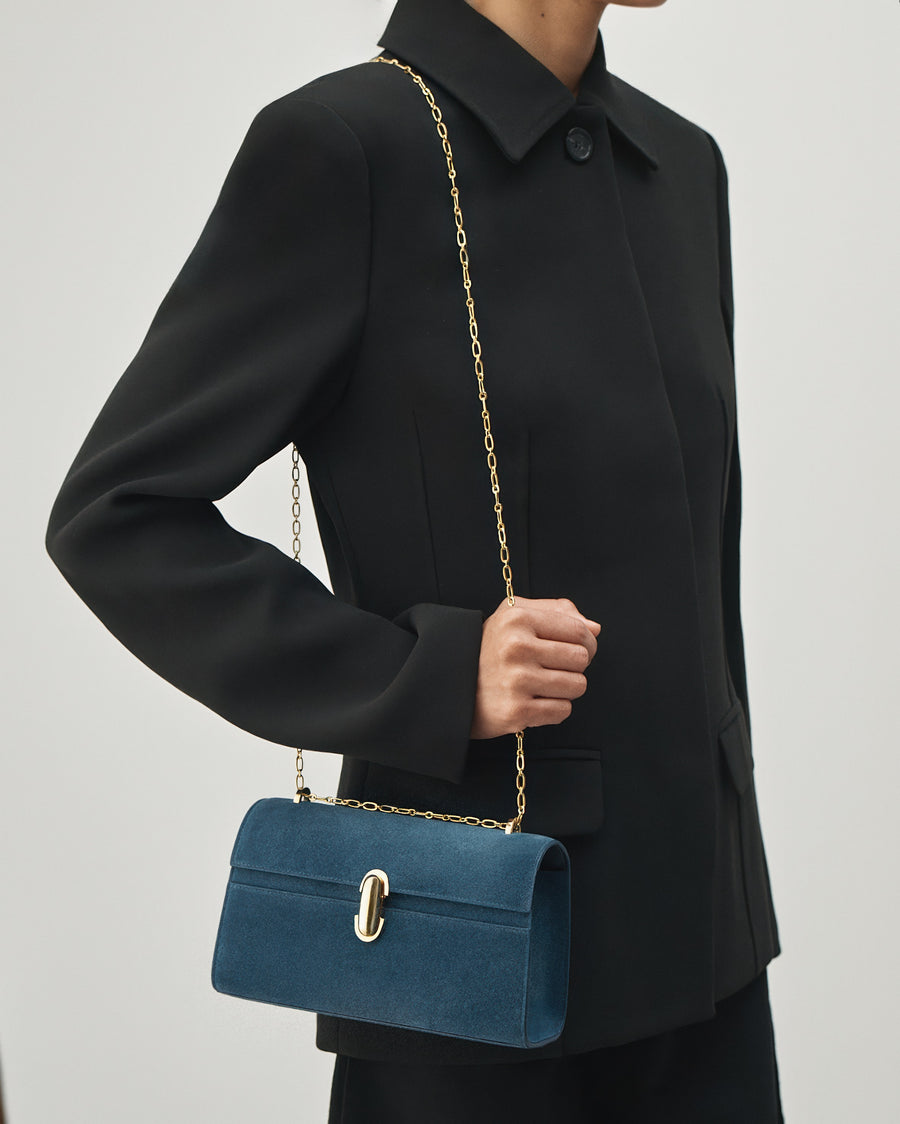 on-model view of a baltic blue suede handbag with a gold clasp and gold chain on a white background 