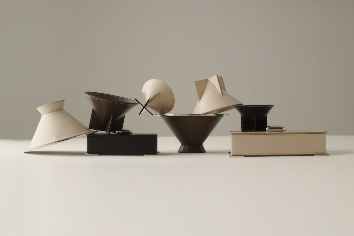 campaign image of stacked mink brown, ebony black, and parchment beige leather dishes and boxes on a grey background
