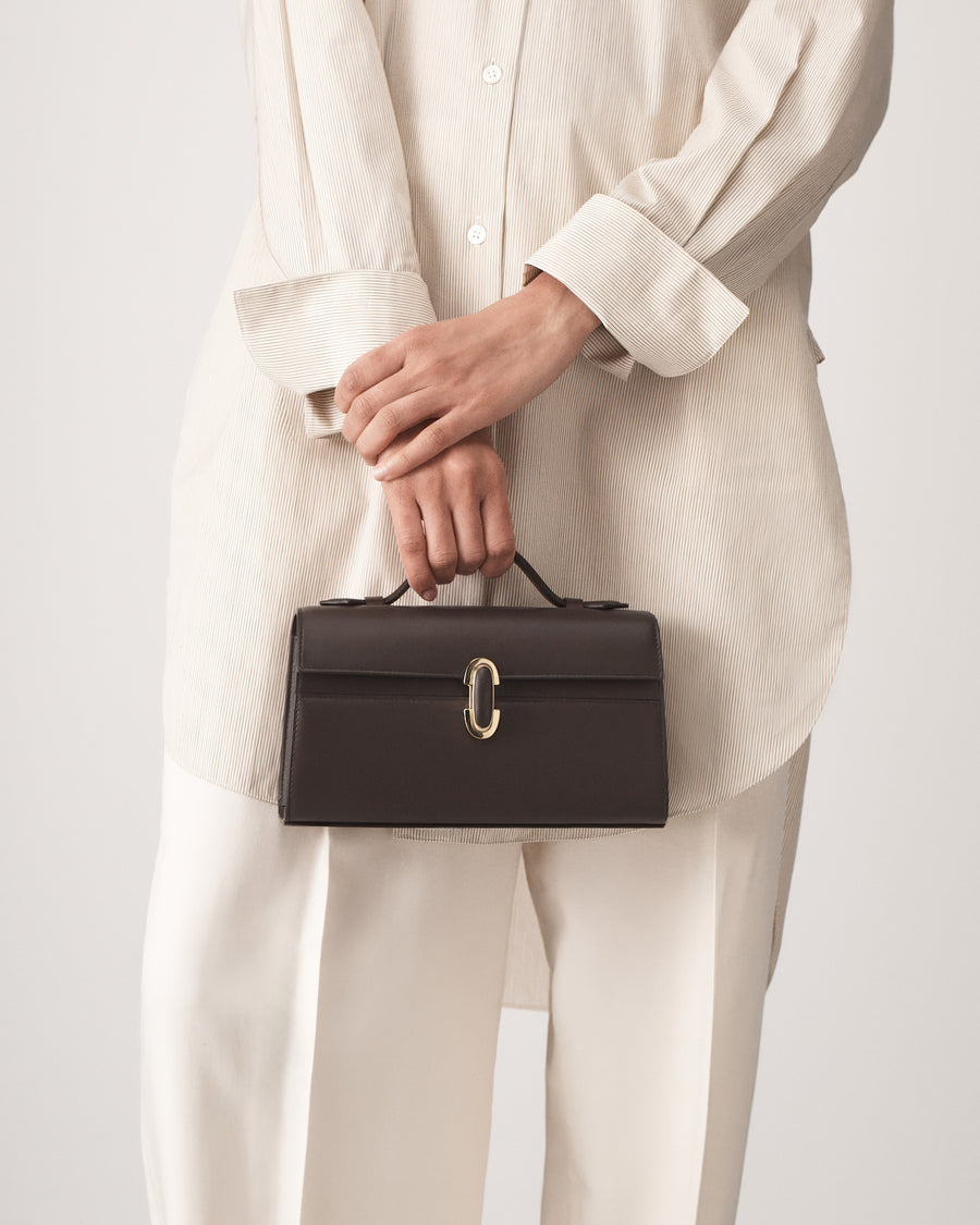 on model view of a coffee brown leather top handle bag with a coffee brown leather covered gold clasp on a white background