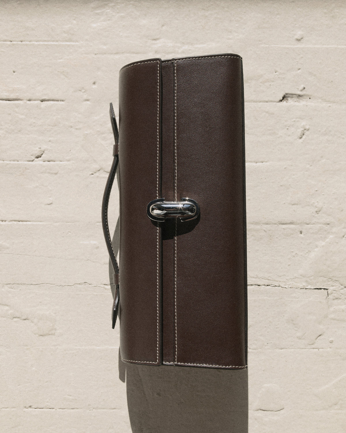 campaign still life image of a currant brown bag with white stitching and silver clasp on a white brick background