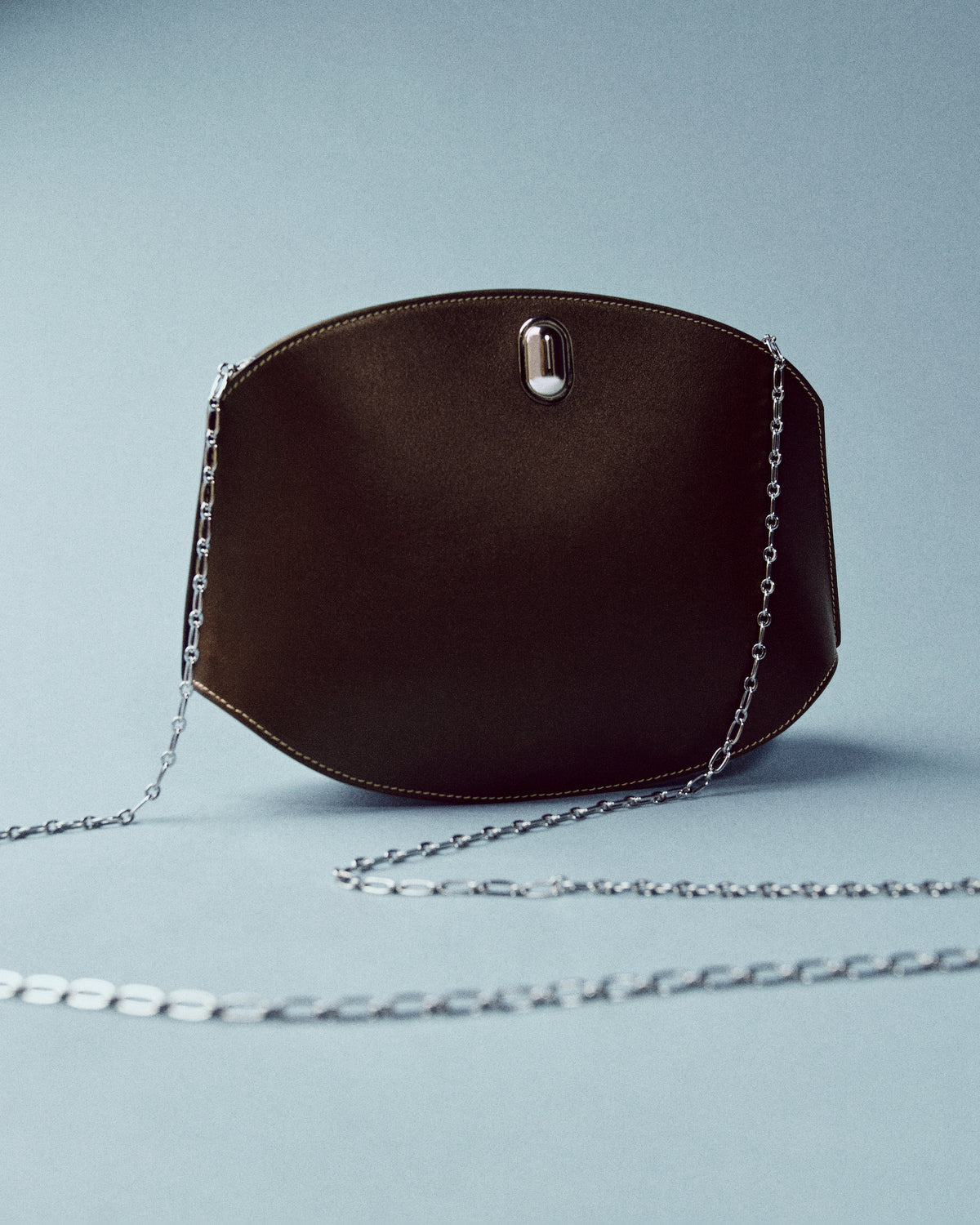 campaign still life image of a brown bag with white stitching and silver hardware with silver chain on a blue background
