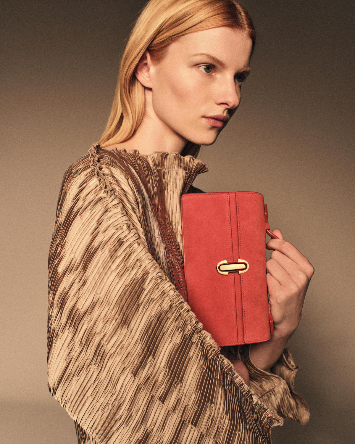 campaign on model image of a poppy red suede bag with a gold clasp on a brown background