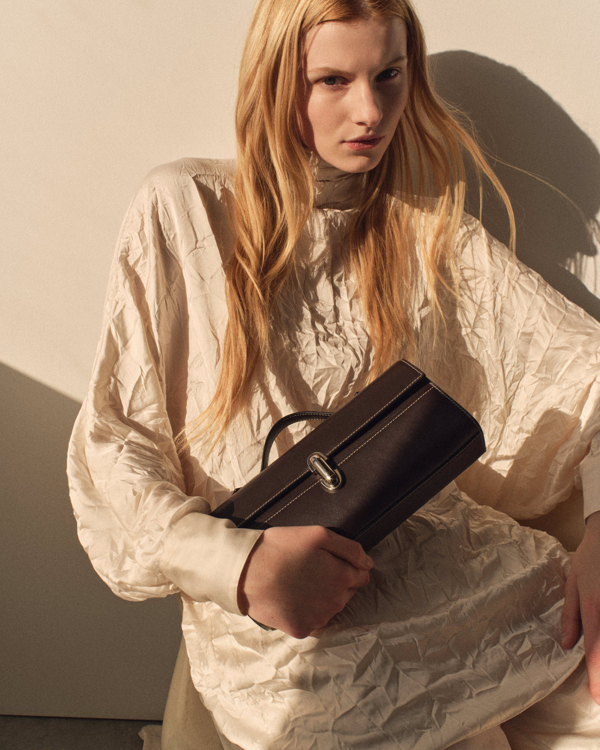 campaign on model image of a currant brown leather bag with a silver clasp on a beige background