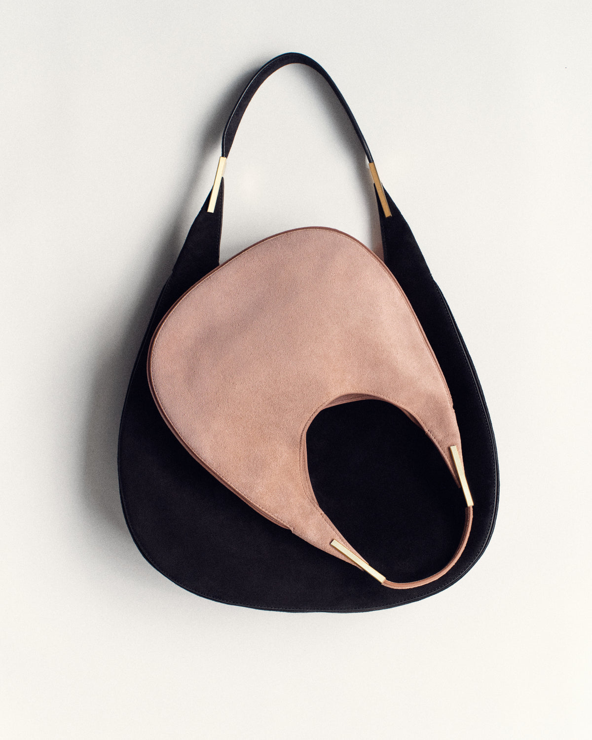 campaign image of a blush pink suede bag with gold hardware on top of a black suede bag with gold hardware on a white background