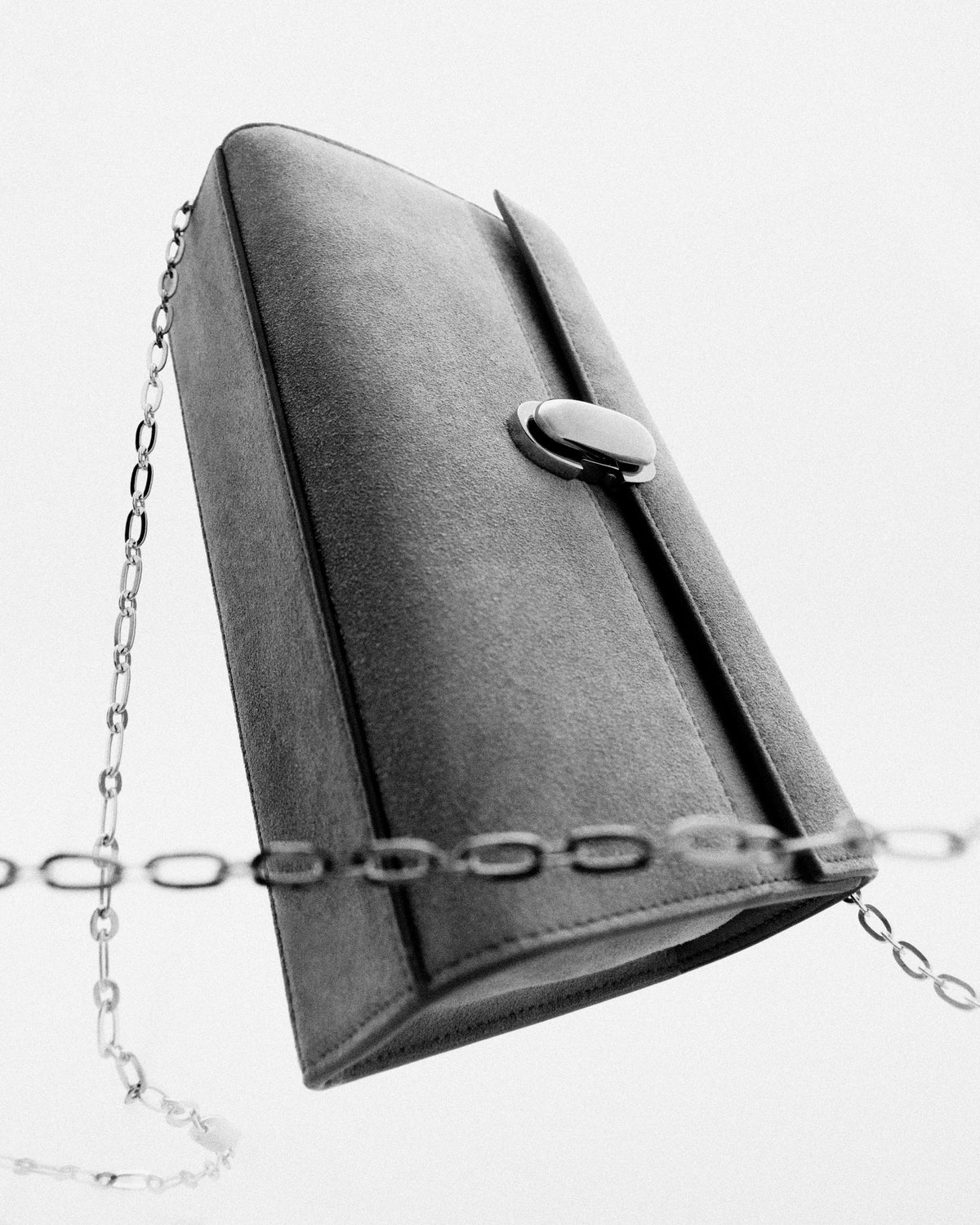 black and white campaign image of a suede bag with metal clasp and chain on a white background