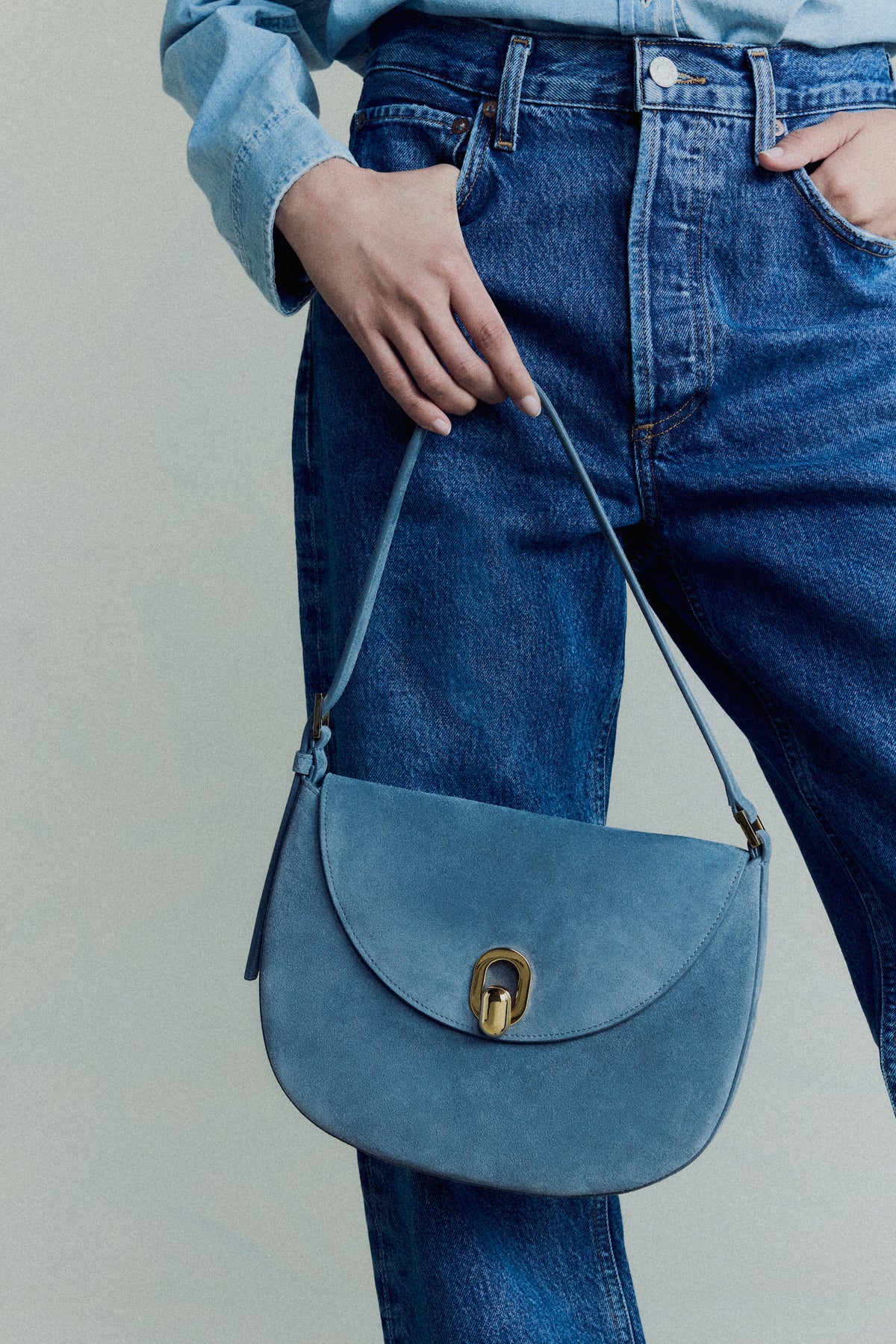 campaign image of a denim blue suede bag with gold hardware on an off white background