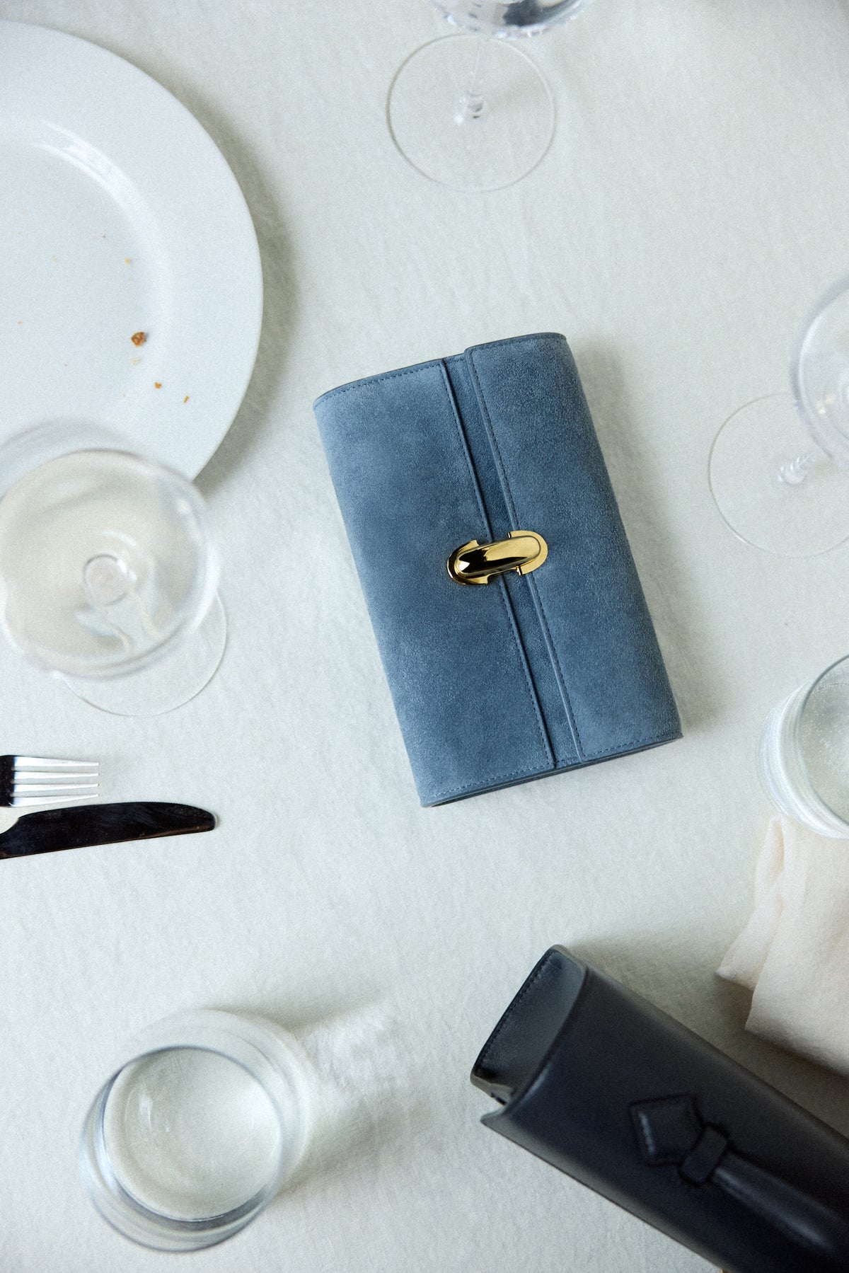 campaign image of a denim blue suede bag with a gold clasp on a dining table background