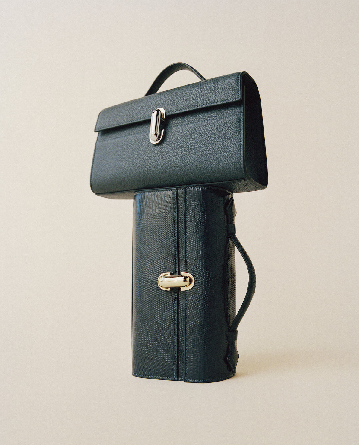 campaign image of black grained leather bag with gold clasp on top of a black lizard back with gold clasp on a beige background