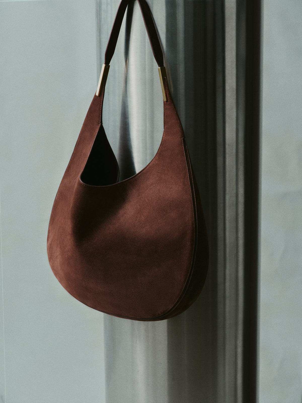 campaign image of espresso brown suede bag with gold hardware hanging on a wall