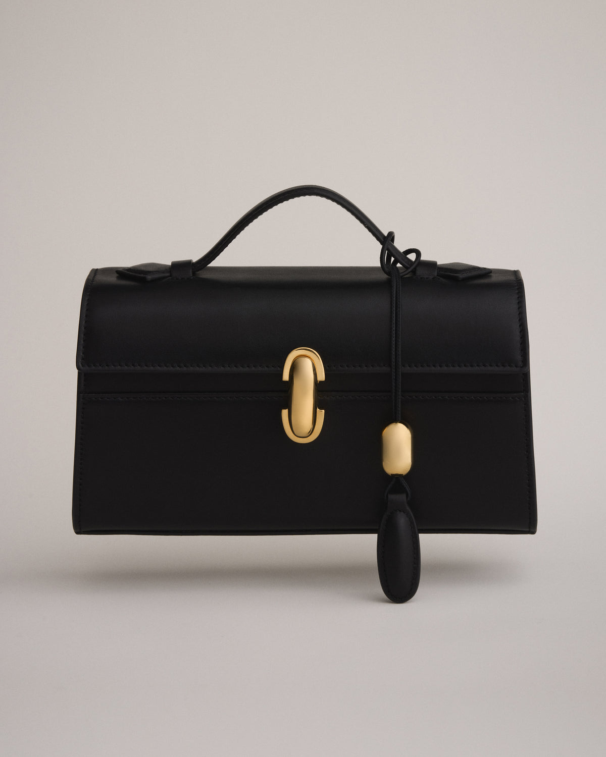 front view of a black leather bag with a gold clasp with a black leather charm on an off white background