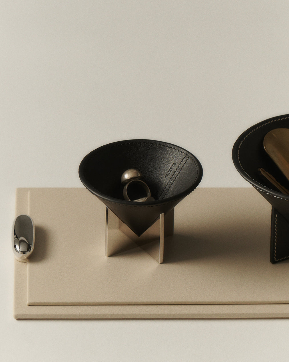 campaign image of a parchment beige leather tray with silver metal hardware with two ebony black leather dishes holding metal jewelry