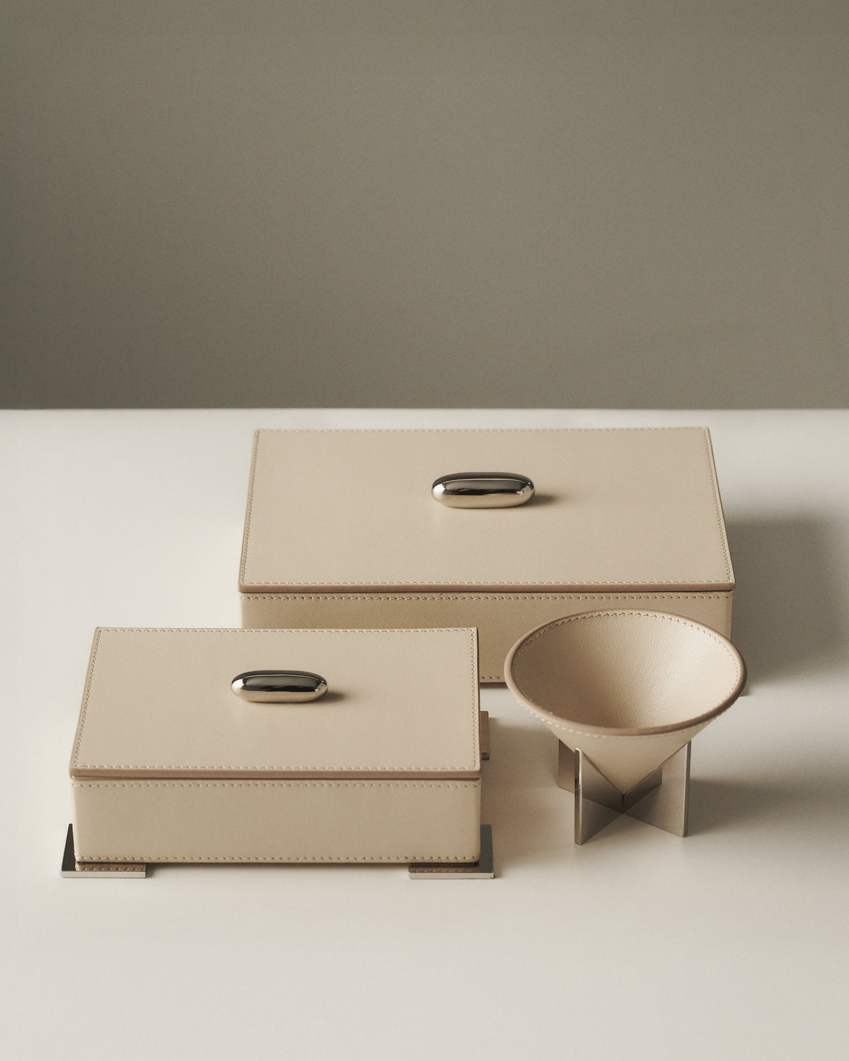 campaign image of two parchment beige leather boxes with silver metal hardware and a small parchment beige leather dish with silver metal feet on a grey background